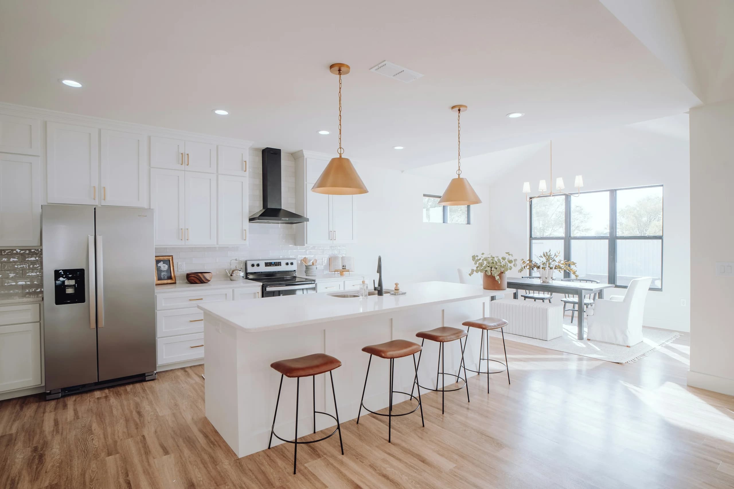 Bright kitchen and dining room with a white island, four brown stools, pale cabinetry and dining seating beside large windows