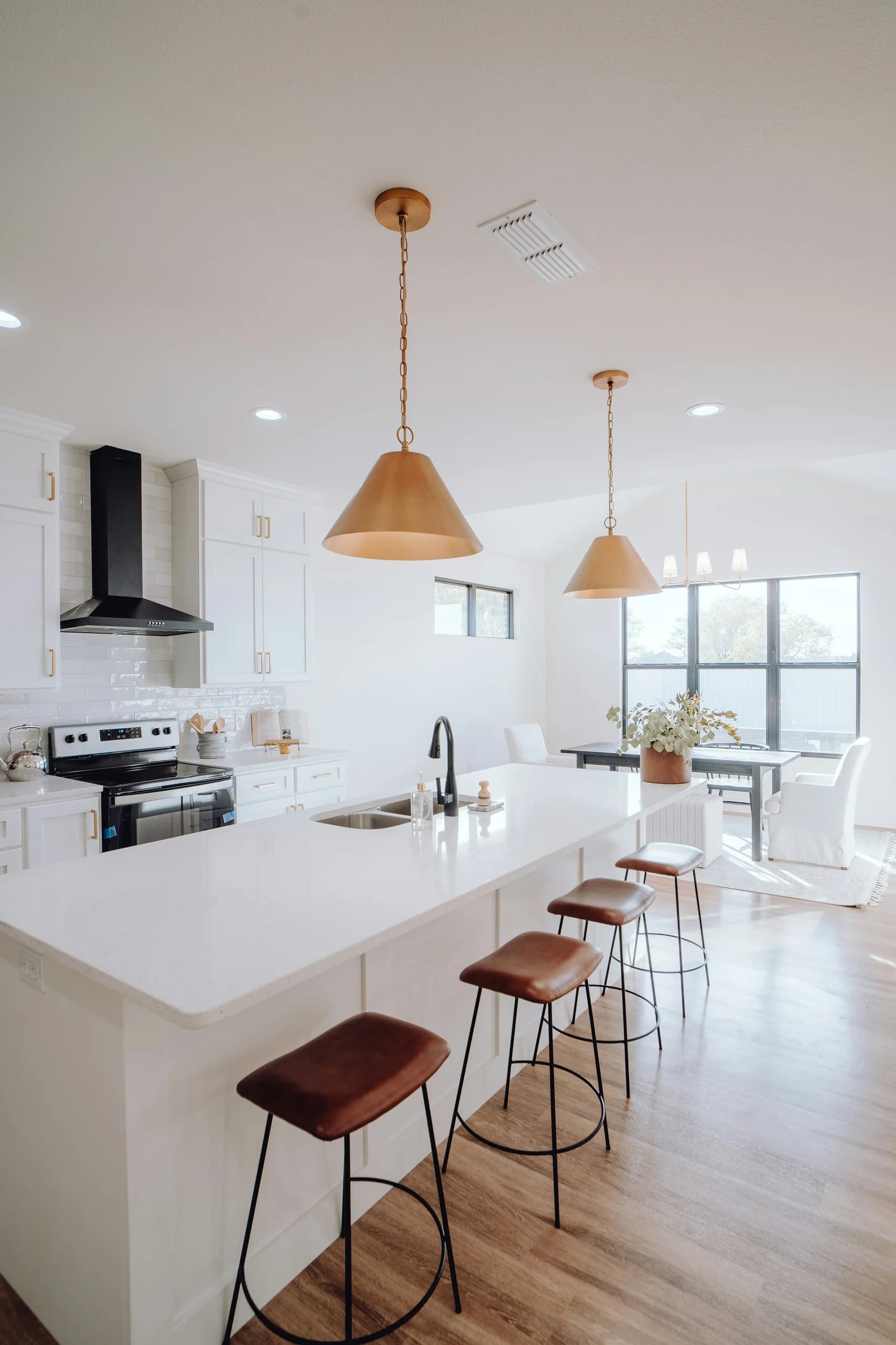 Portrait view along a long white kitchen island with brown stools, warm metal pendants, a sink and dining seating beyond