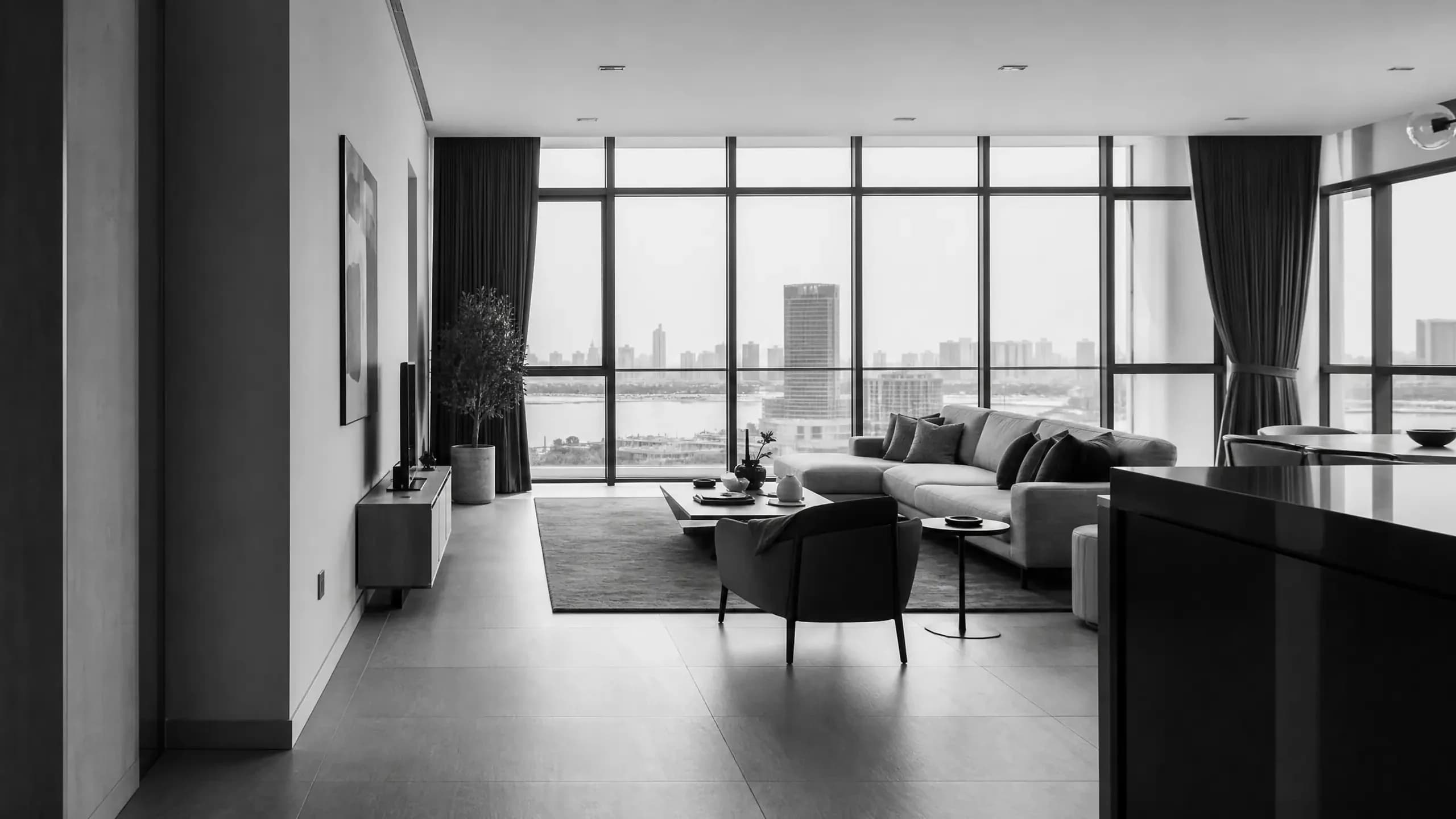Monochrome view of a living room with a long sofa, an armchair, a media console and floor-to-ceiling city windows