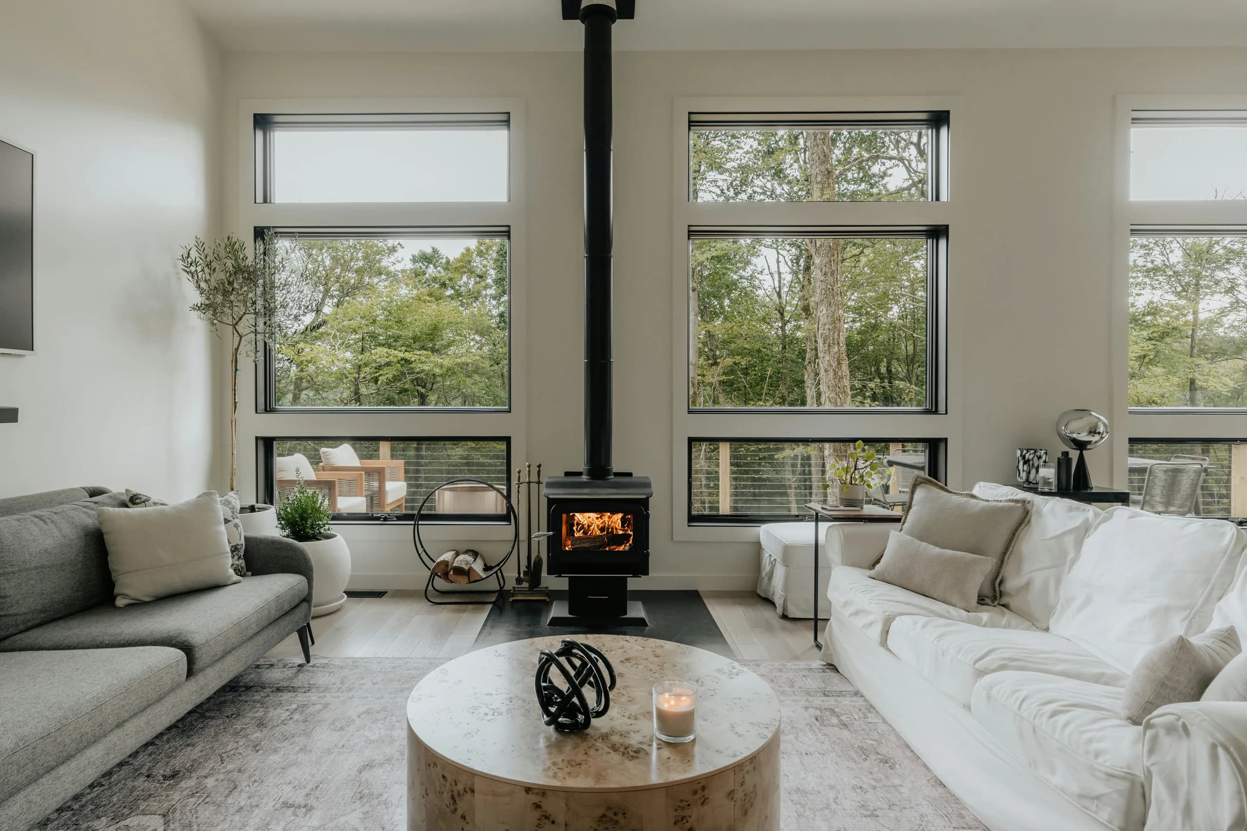 Grey and white sofas facing a round pale table, with tall woodland-facing windows and a central black stove
